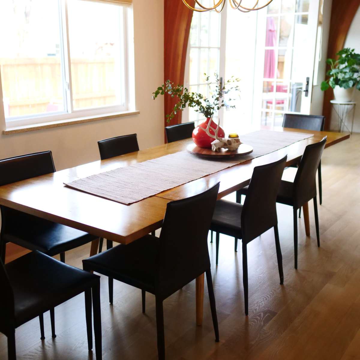 A long wooden dining table is surrounded by eight black chairs in a well-lit room with large windows.