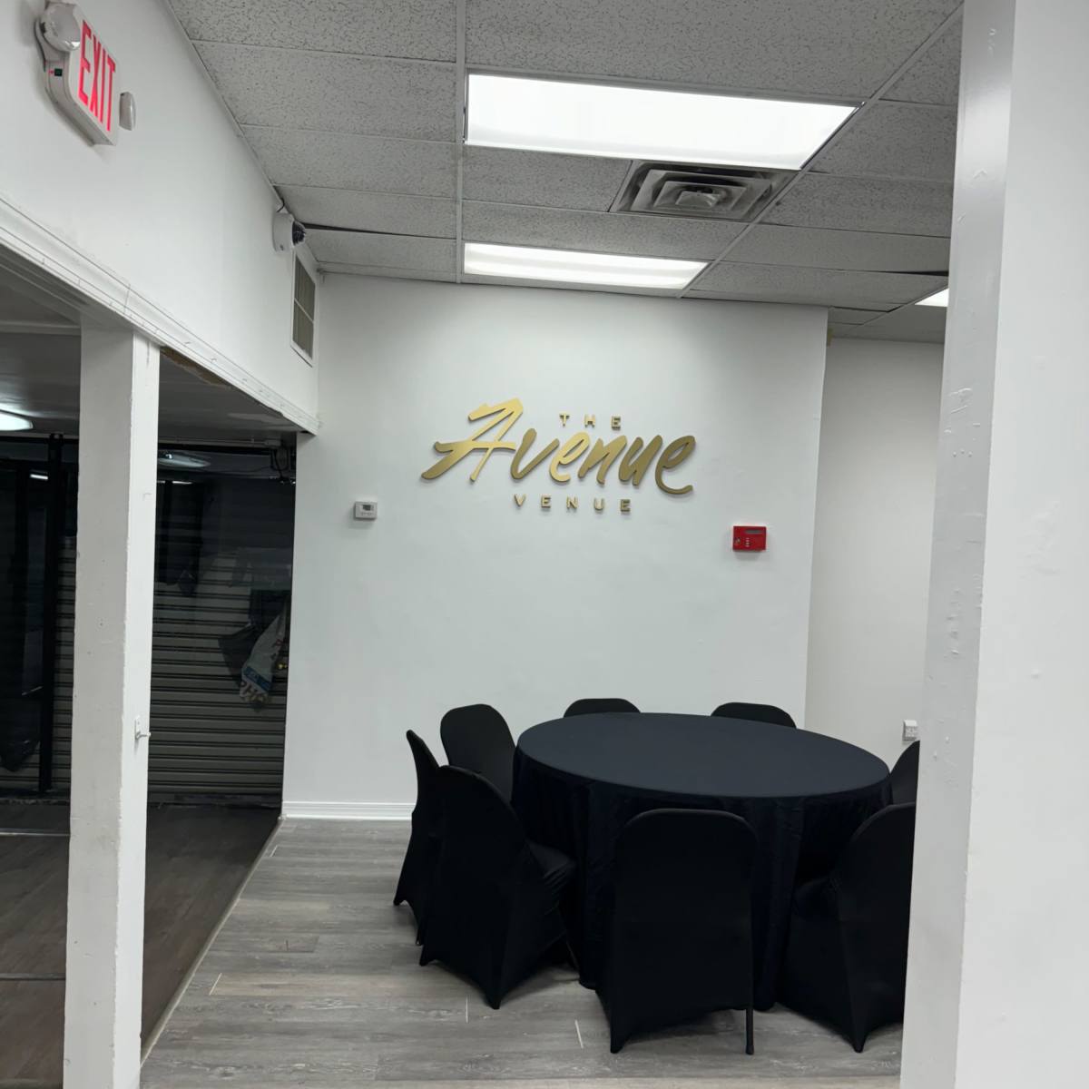 A spacious indoor venue with a circular black table surrounded by black chairs, and a gold sign on the wall reading "The Avenue Venue."