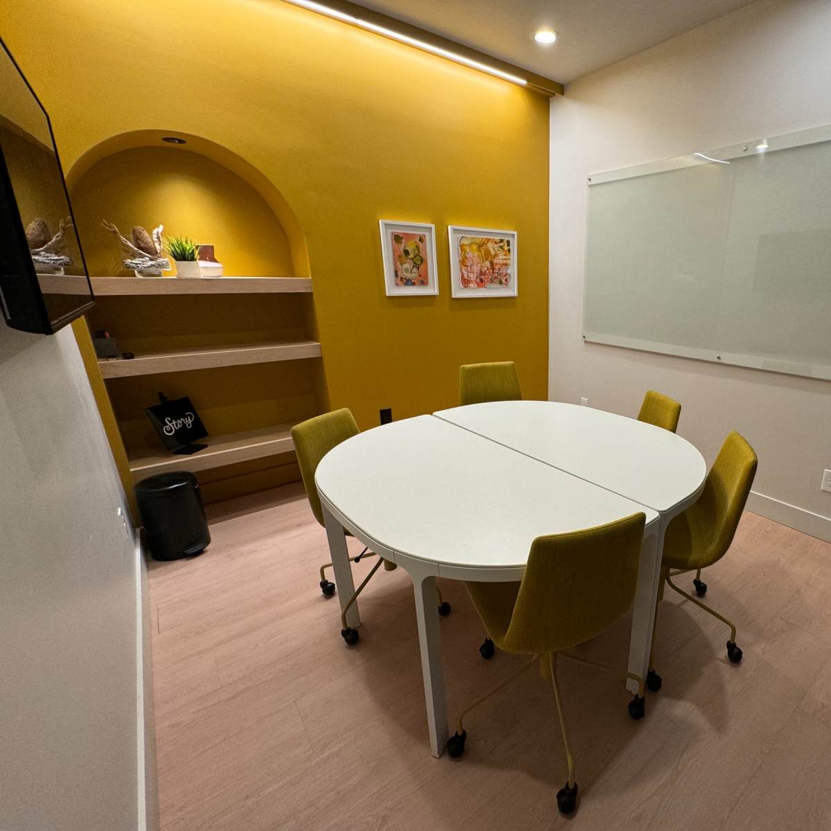 A small meeting room features a round white table surrounded by four yellow chairs, with a light yellow wall and a glass whiteboard.