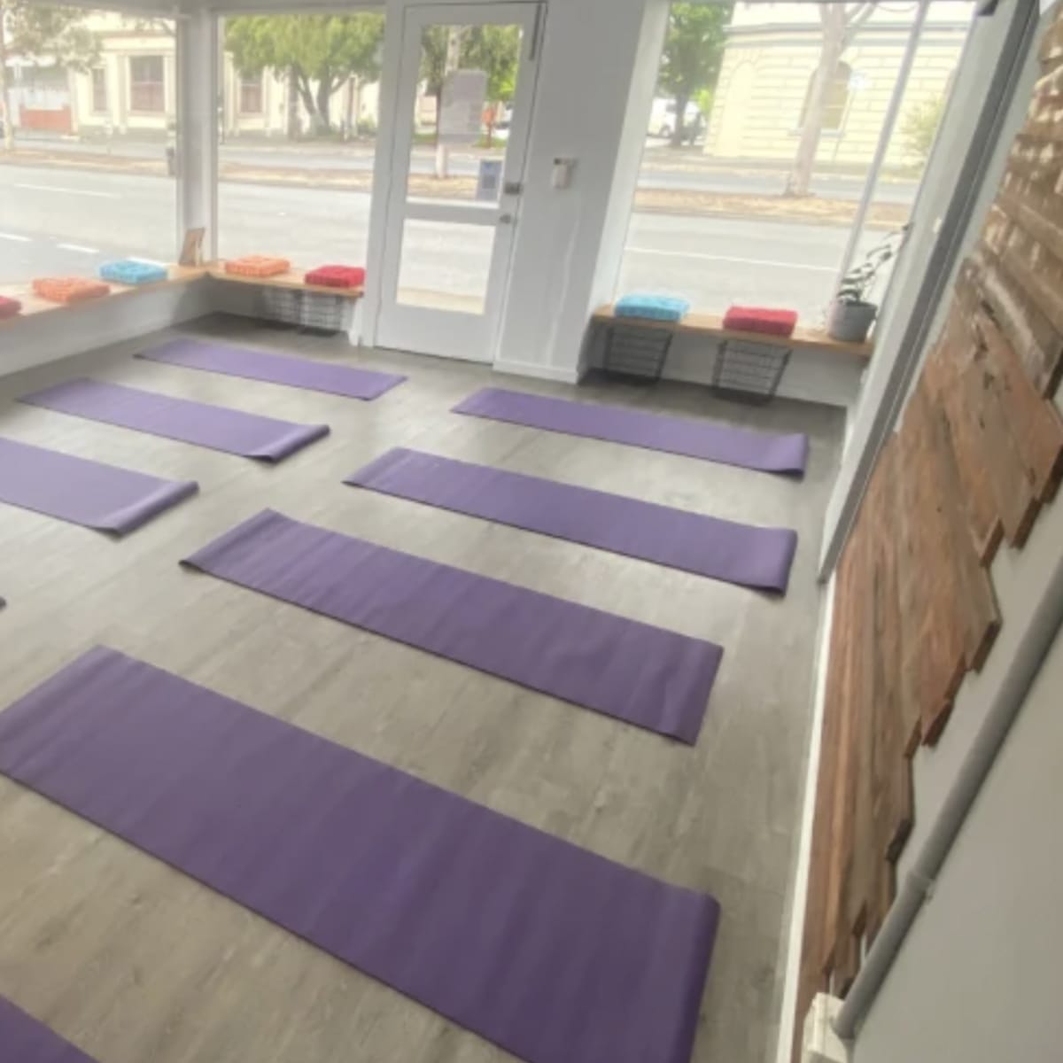 A yoga studio with several purple mats arranged in rows on a wooden floor, and colorful cushions lined up on a window ledge.