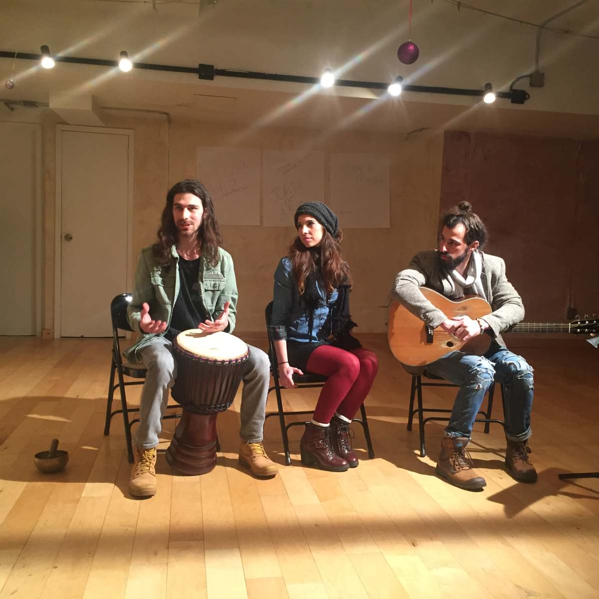 Three musicians sit in chairs in a well-lit room, with one playing a djembe drum, another seated in the middle, and the third holding an acoustic guitar.