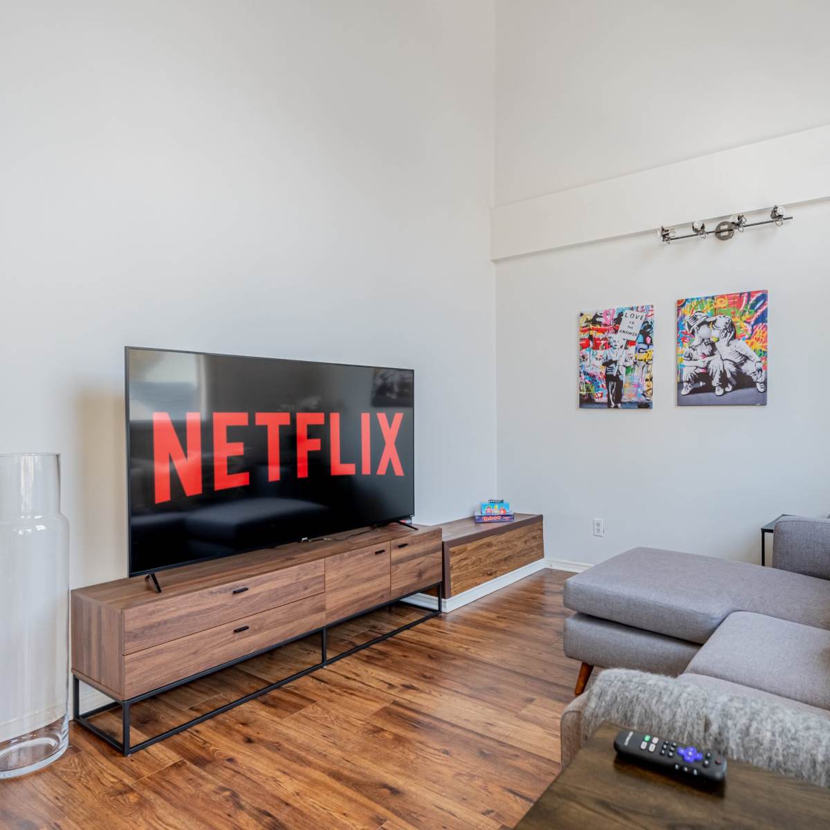 A living room with a large television displaying the Netflix logo, a grey sofa, and two colorful wall art pieces.