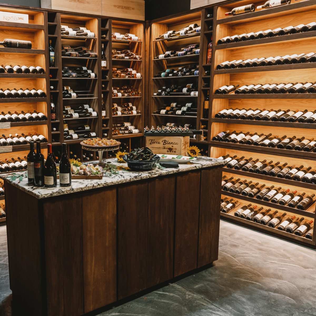 A wine cellar features wooden shelving filled with bottles of wine, with a marble-topped table in the center adorned with snacks and wine bottles.