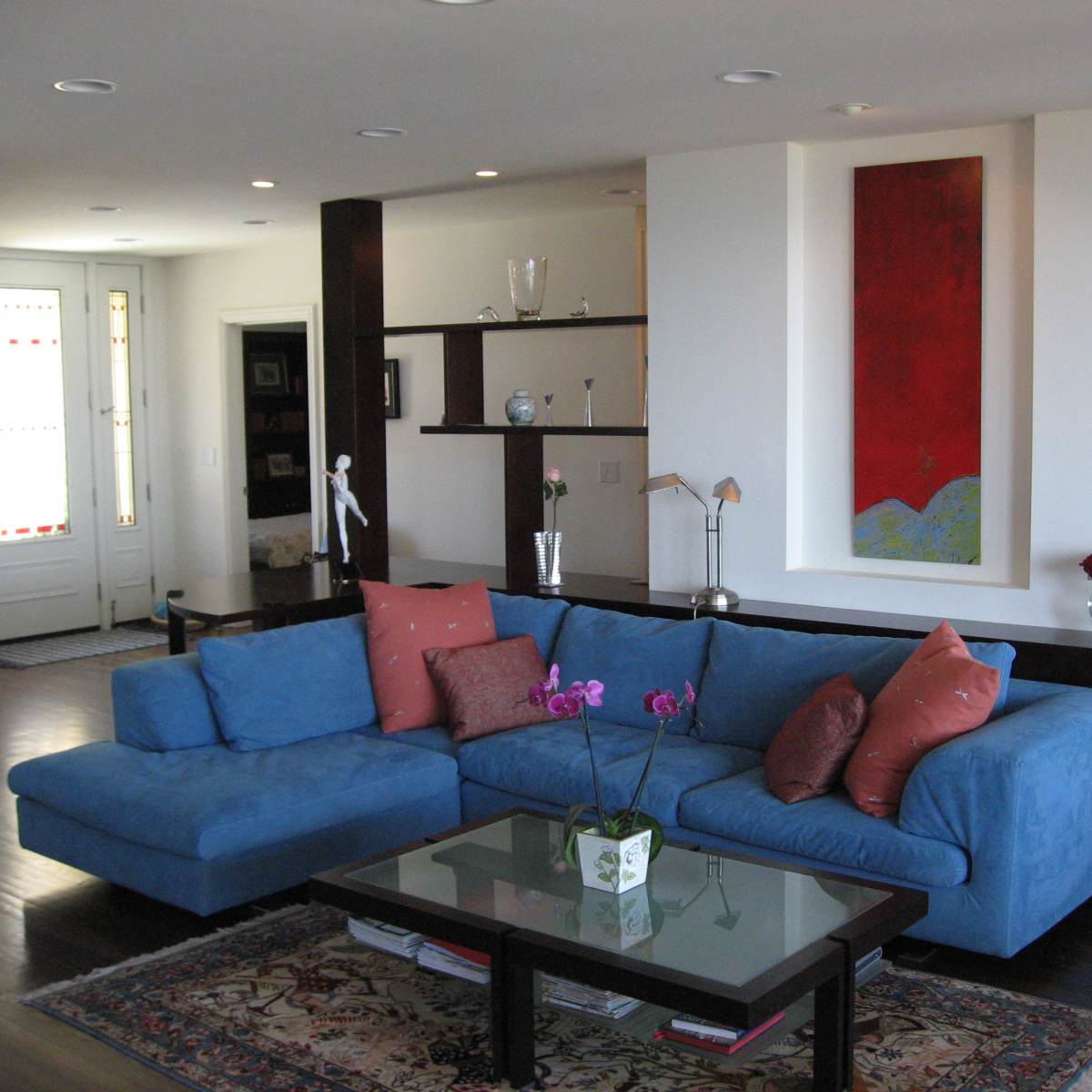 A modern living room featuring a blue sectional couch, a glass coffee table, and a red abstract painting on the wall.