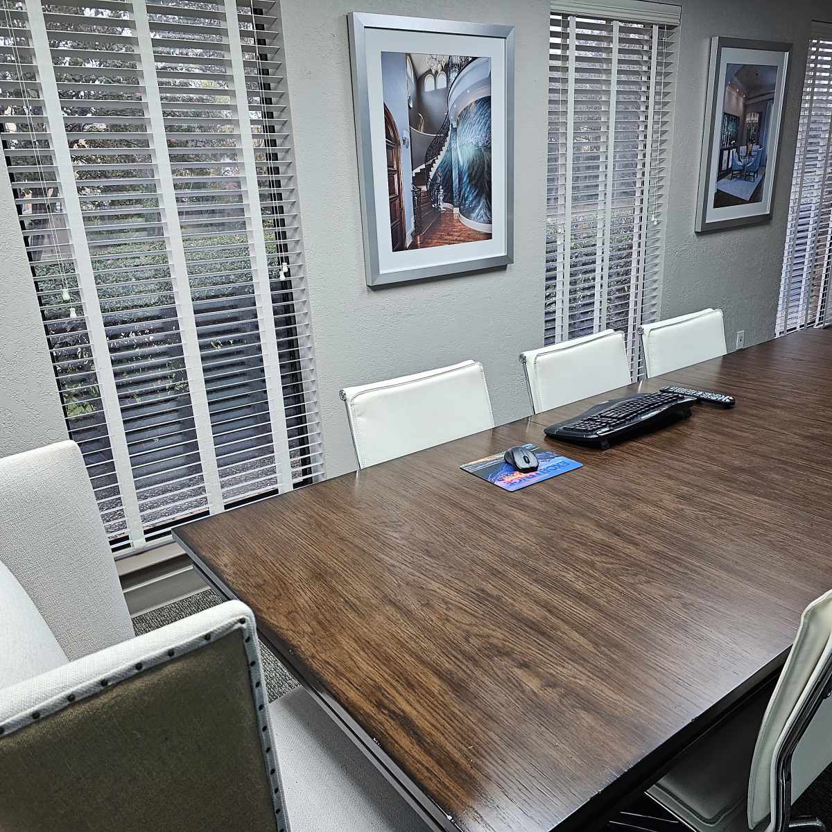A long wooden conference table is surrounded by several white chairs in a well-lit room with large windows covered by blinds.