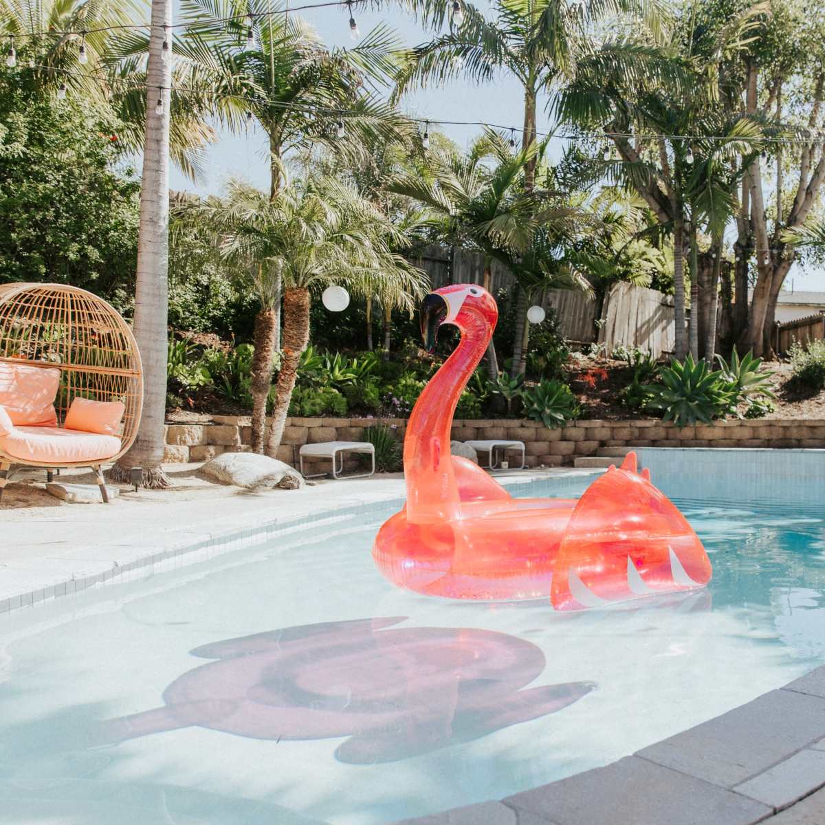 A bright pink flamingo float sits in a swimming pool surrounded by palm trees and landscaped greenery.