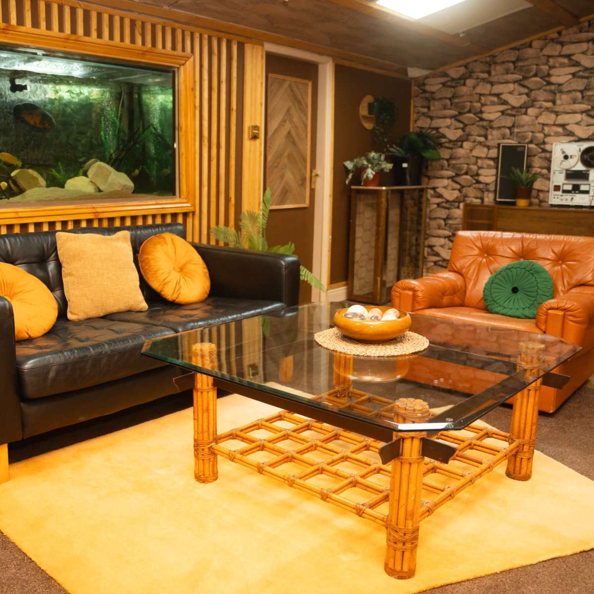 The living room features a black leather sofa, a brown armchair, a glass coffee table with a rattan base, and a large aquarium set against a wooden wall.