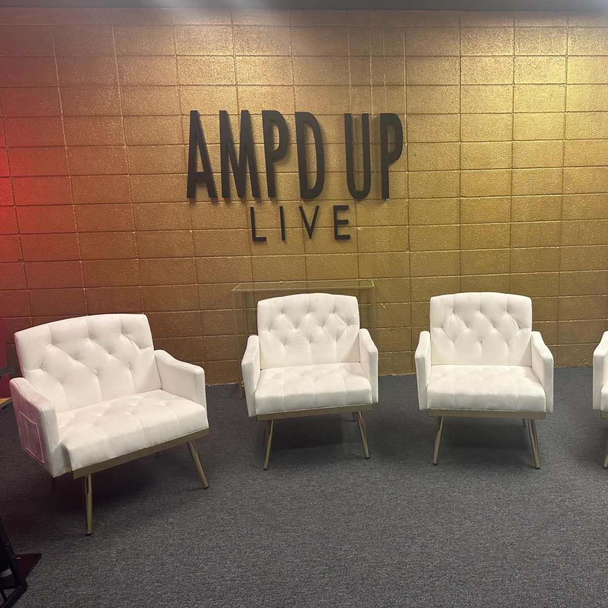 Four white tufted armchairs arranged in front of a gold and black wall with the text "AMPD UP LIVE" displayed.