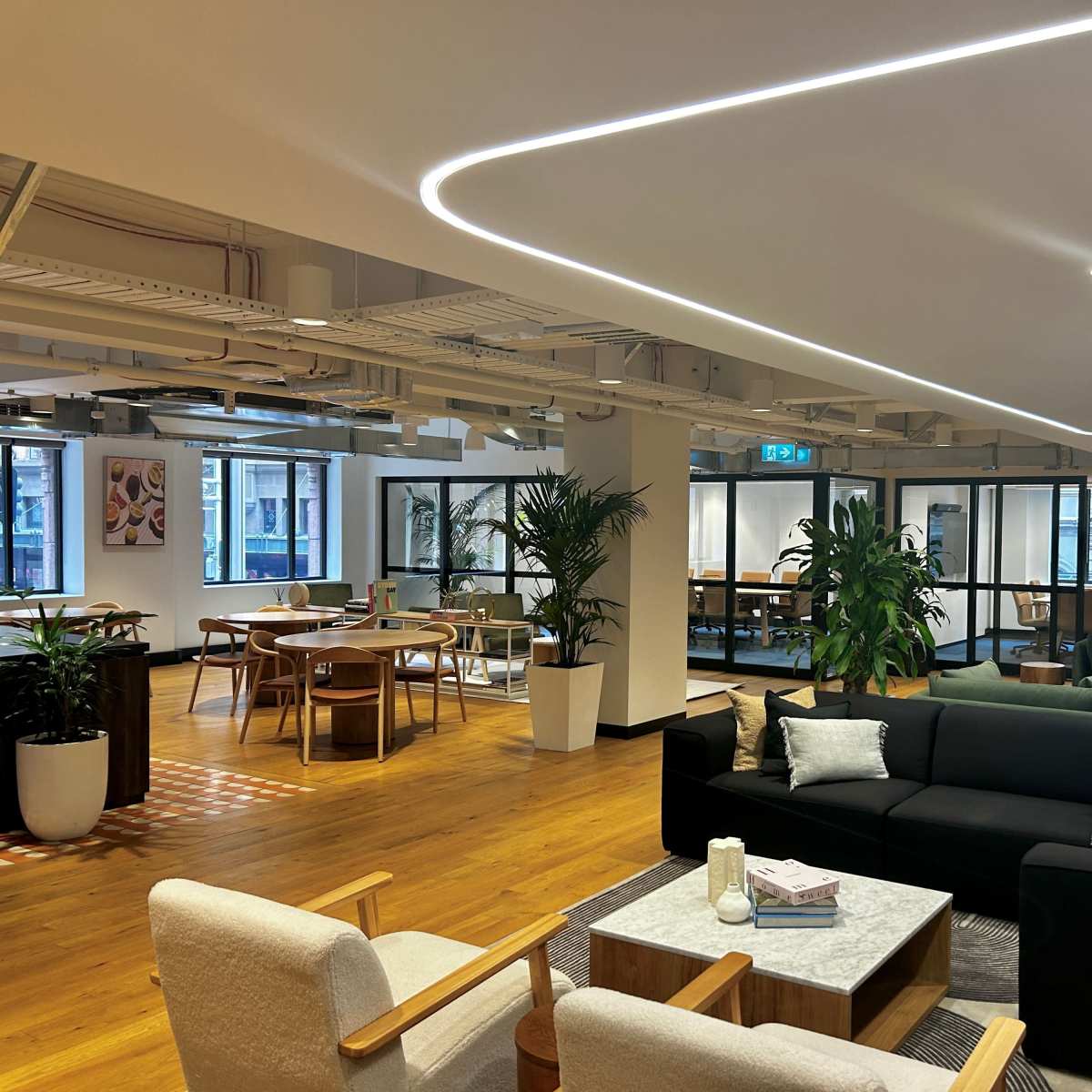 A modern office interior featuring a lounge area with sofas, chairs, plants, and workspaces surrounded by large windows.