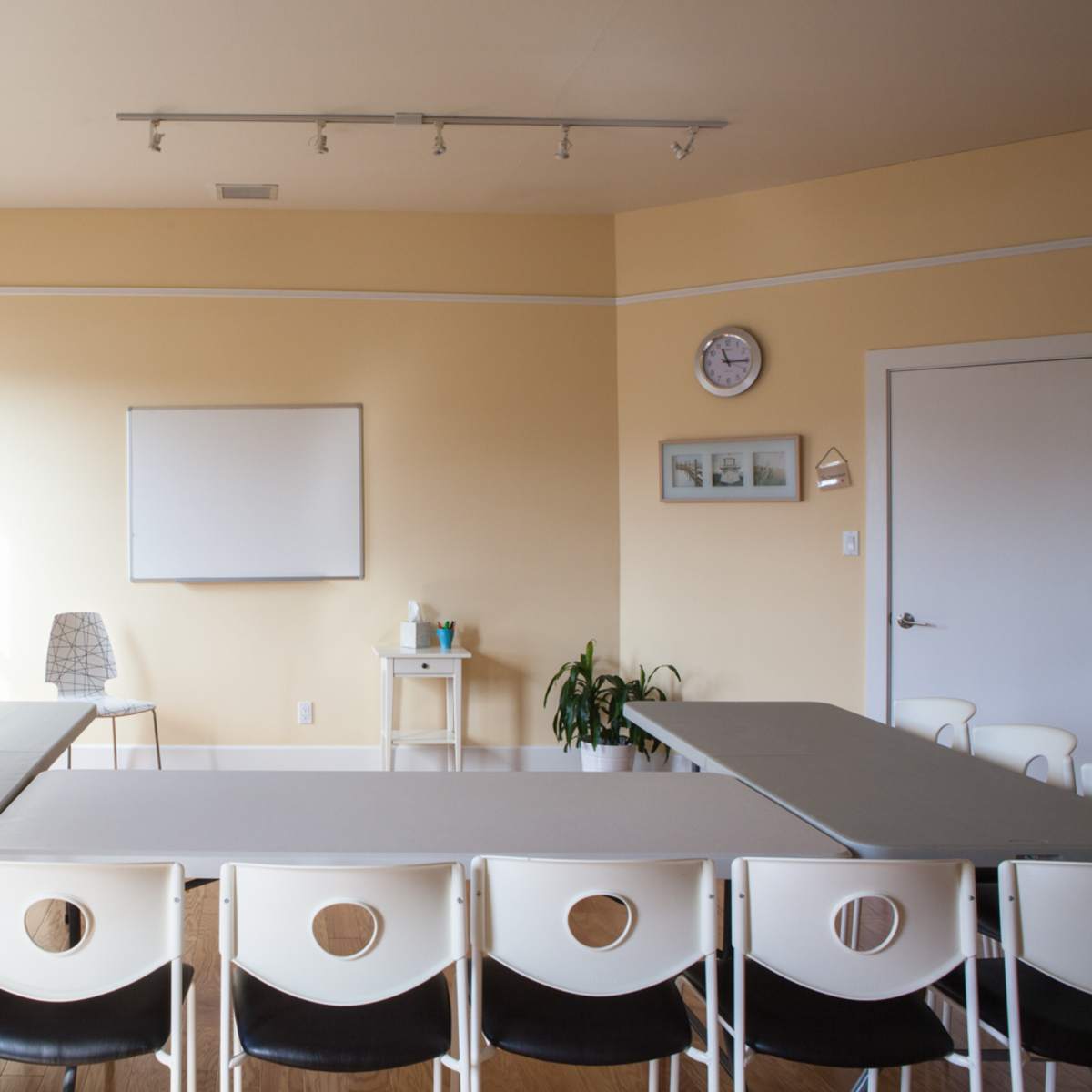 A bright, empty classroom features a rectangular arrangement of tables and chairs, a whiteboard, a clock, and a small plant in the corner.