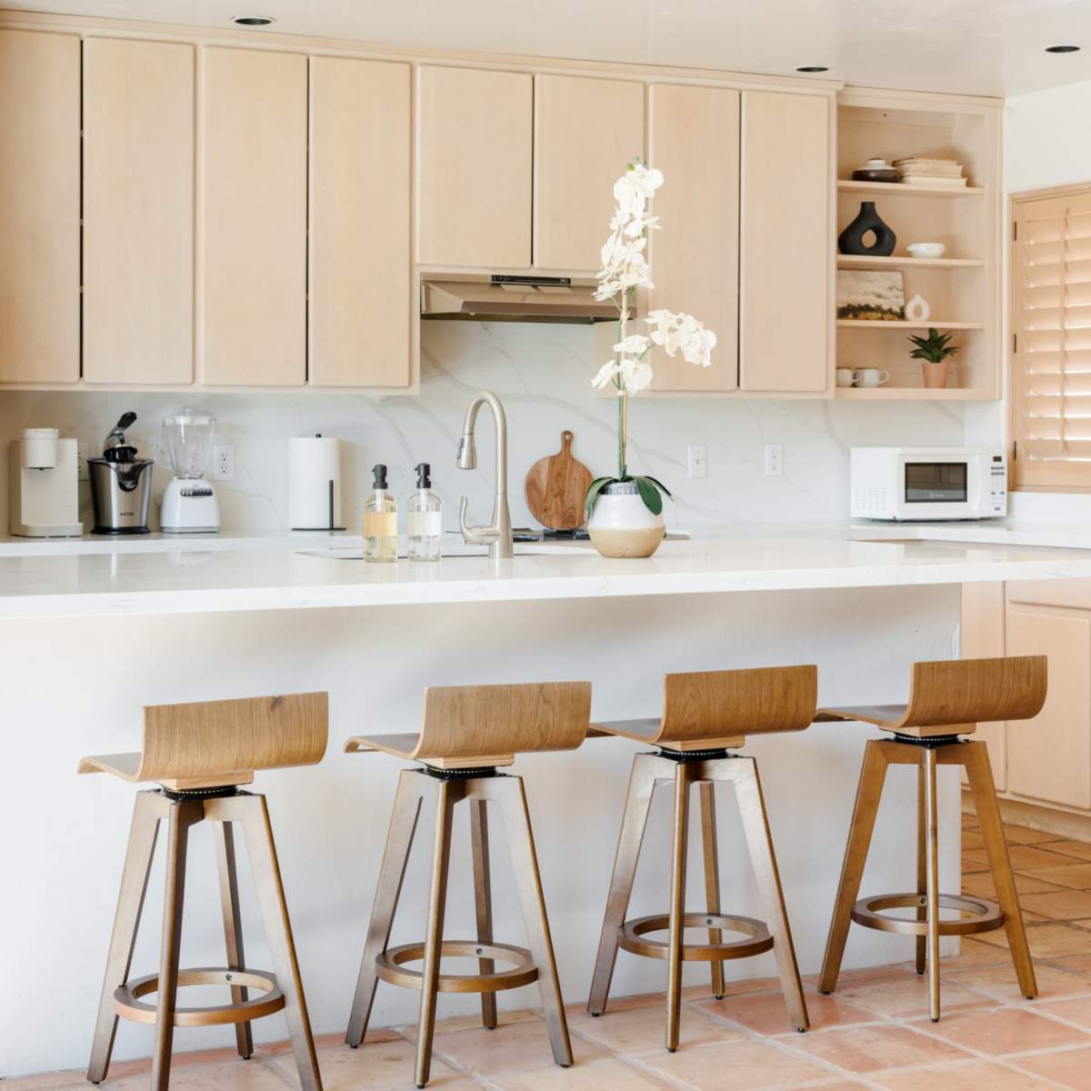A minimalist kitchen features light wooden cabinetry, a countertop with a sink, and four wooden bar stools arranged in front.