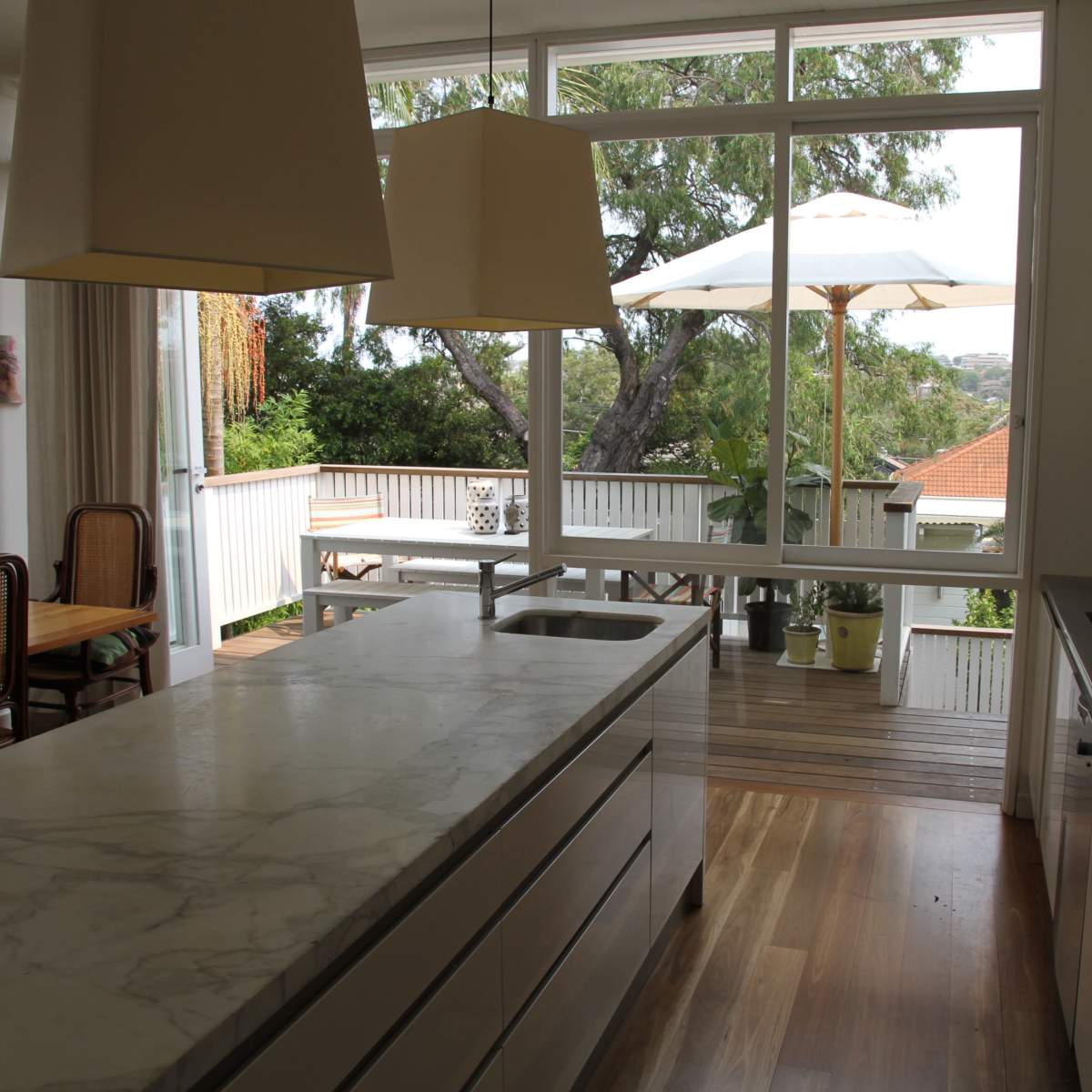A modern kitchen featuring a large marble island and open windows that overlook a deck with outdoor seating and an umbrella.