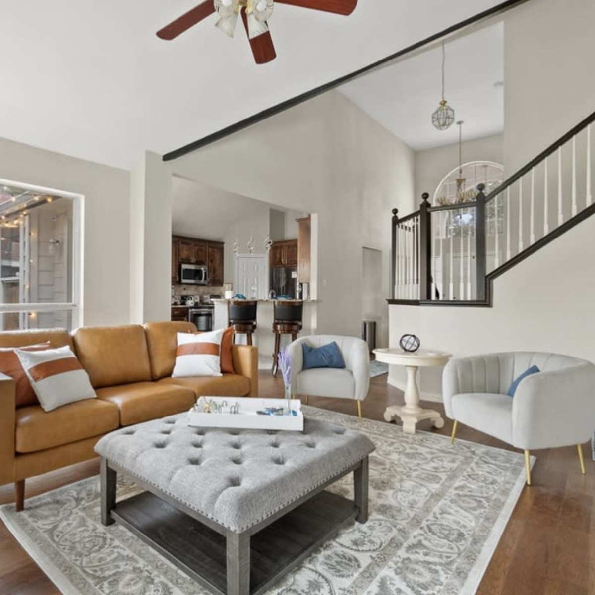 A modern living room features a brown leather sofa, a gray tufted coffee table, and a view of a staircase leading to an upper level.