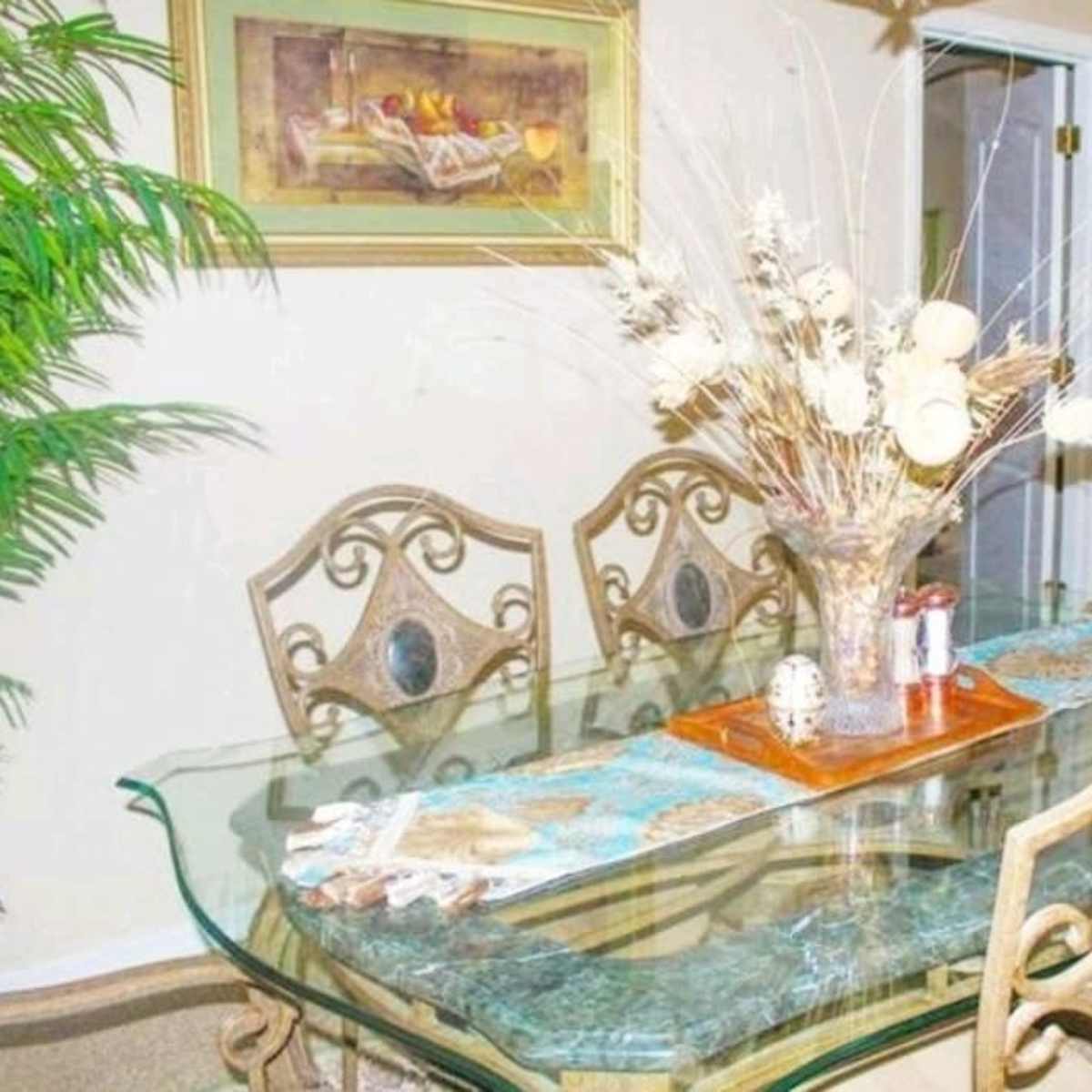 A glass dining table is set with a decorative centerpiece and surrounded by ornate metal chairs in a well-lit room.