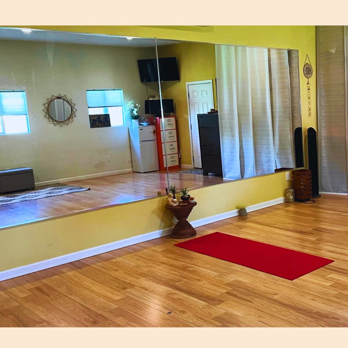 A spacious room with wooden flooring, a large mirror on one wall, a red yoga mat on the floor, and a wooden dresser near a door.