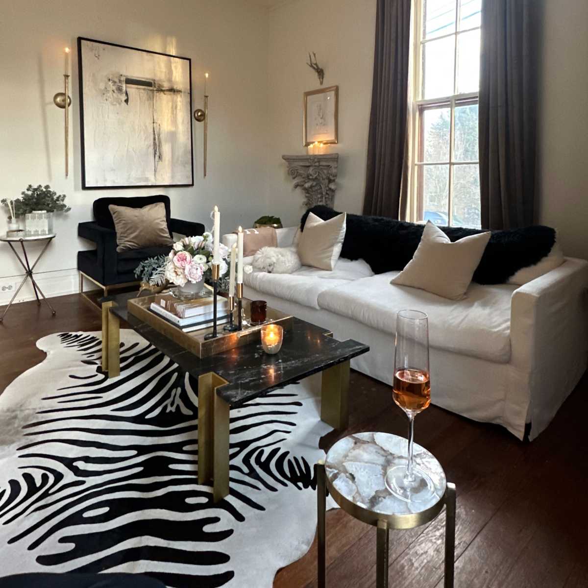The living room features a white sofa, a black armchair, a zebra-patterned rug, and a coffee table adorned with candles and floral arrangements under a chandelier.