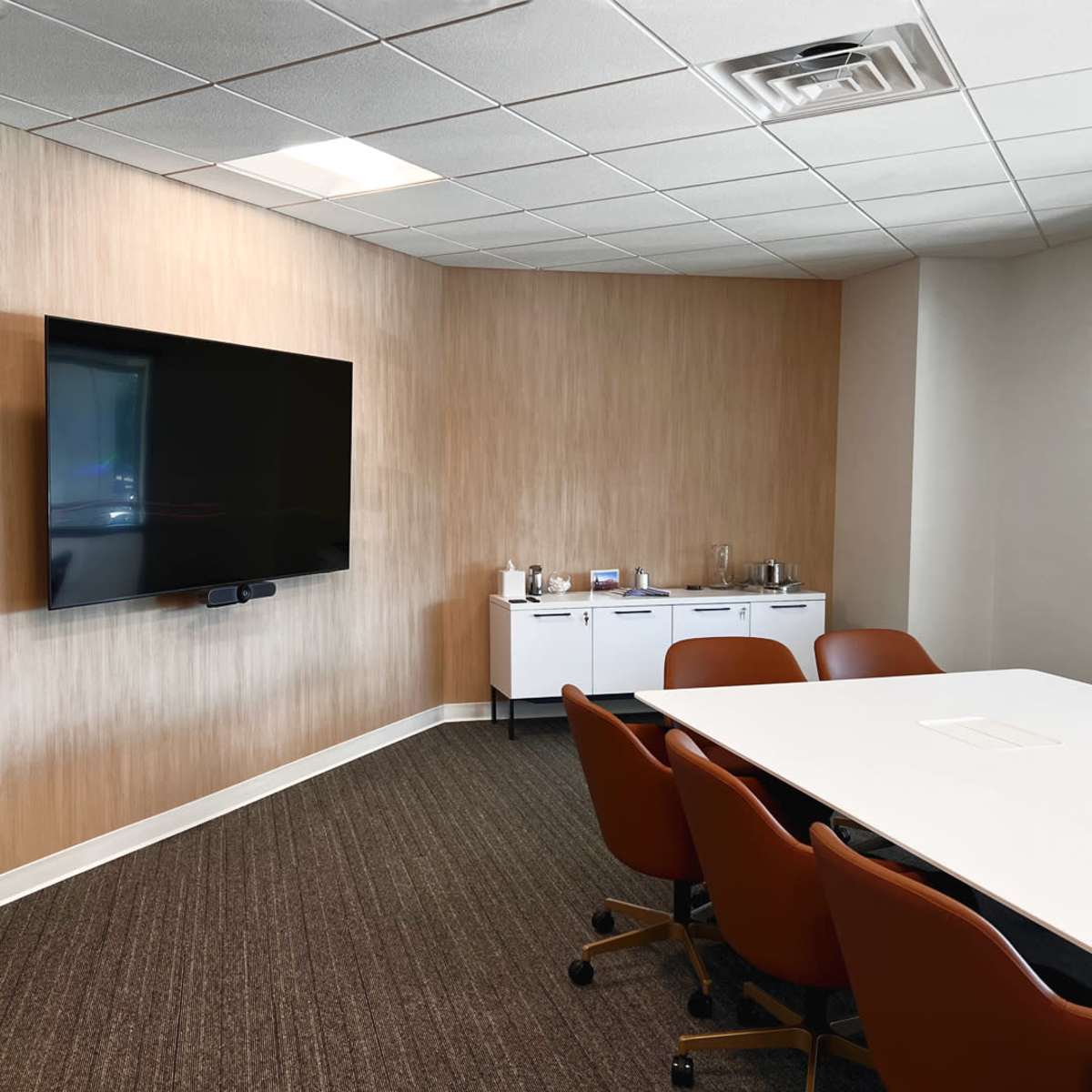 A modern meeting room with a large wall-mounted TV, a white conference table, and brown chairs.