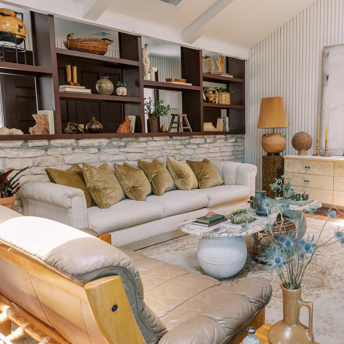 A cozy living room featuring a light-colored sofa with multiple cushions, a wicker chair, a wooden side table, and a stone wall bookshelf filled with decorative items.
