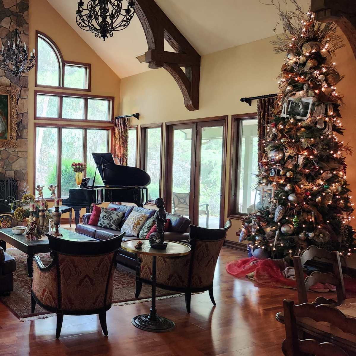 A spacious living room with a tall stone fireplace, large windows, a grand piano, and a decorated Christmas tree in the corner.