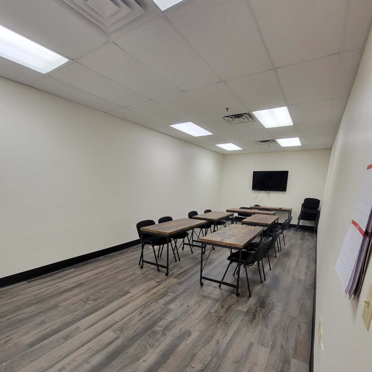 A sparsely furnished meeting room with a rectangular table, black chairs, and a wall-mounted screen.