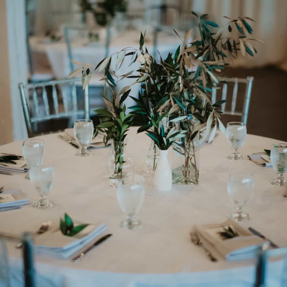 A round table is set with water glasses, napkins, and a centerpiece of greenery in a decorated venue.