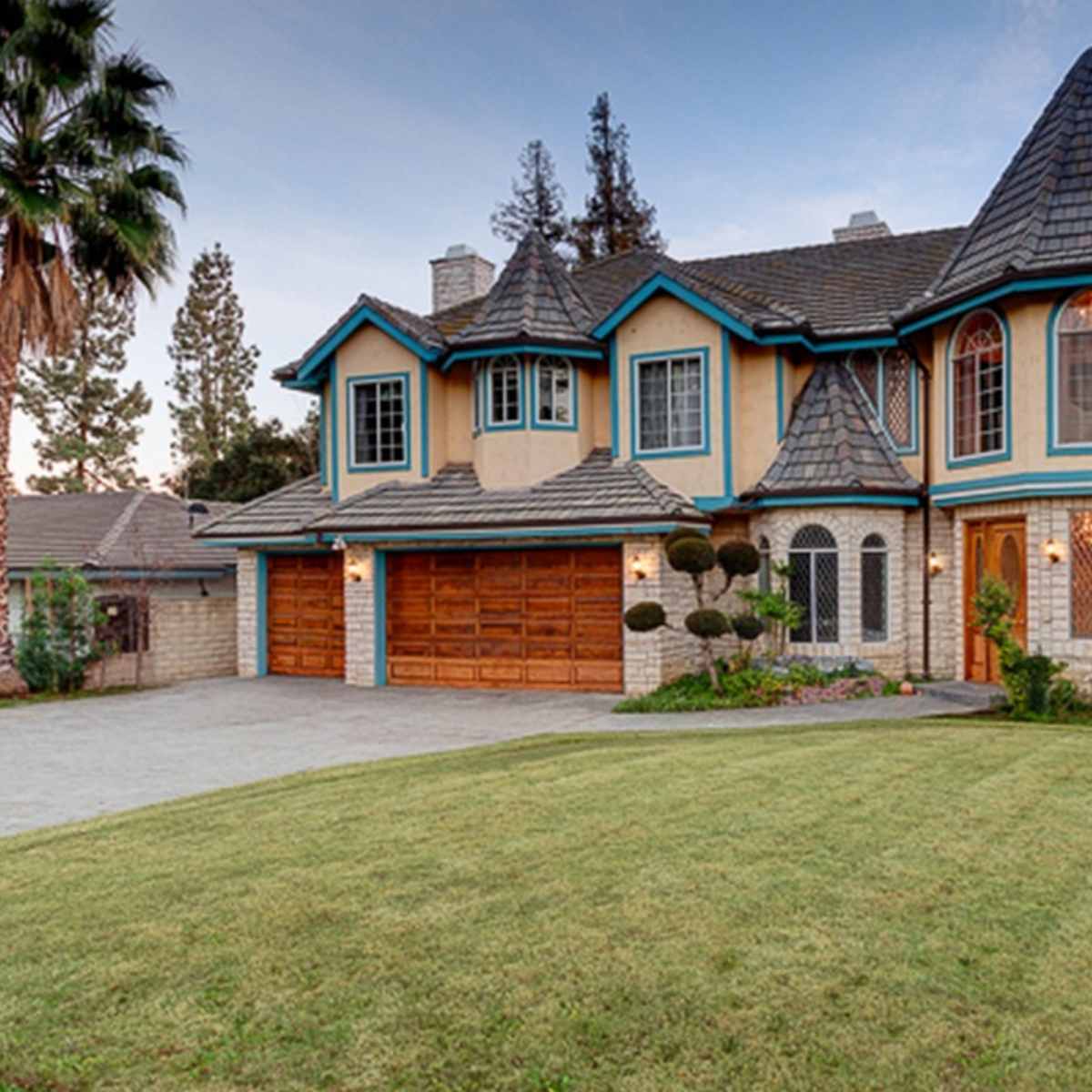 A large, two-story house with a turret and a stone facade, surrounded by manicured lawns and palm trees.