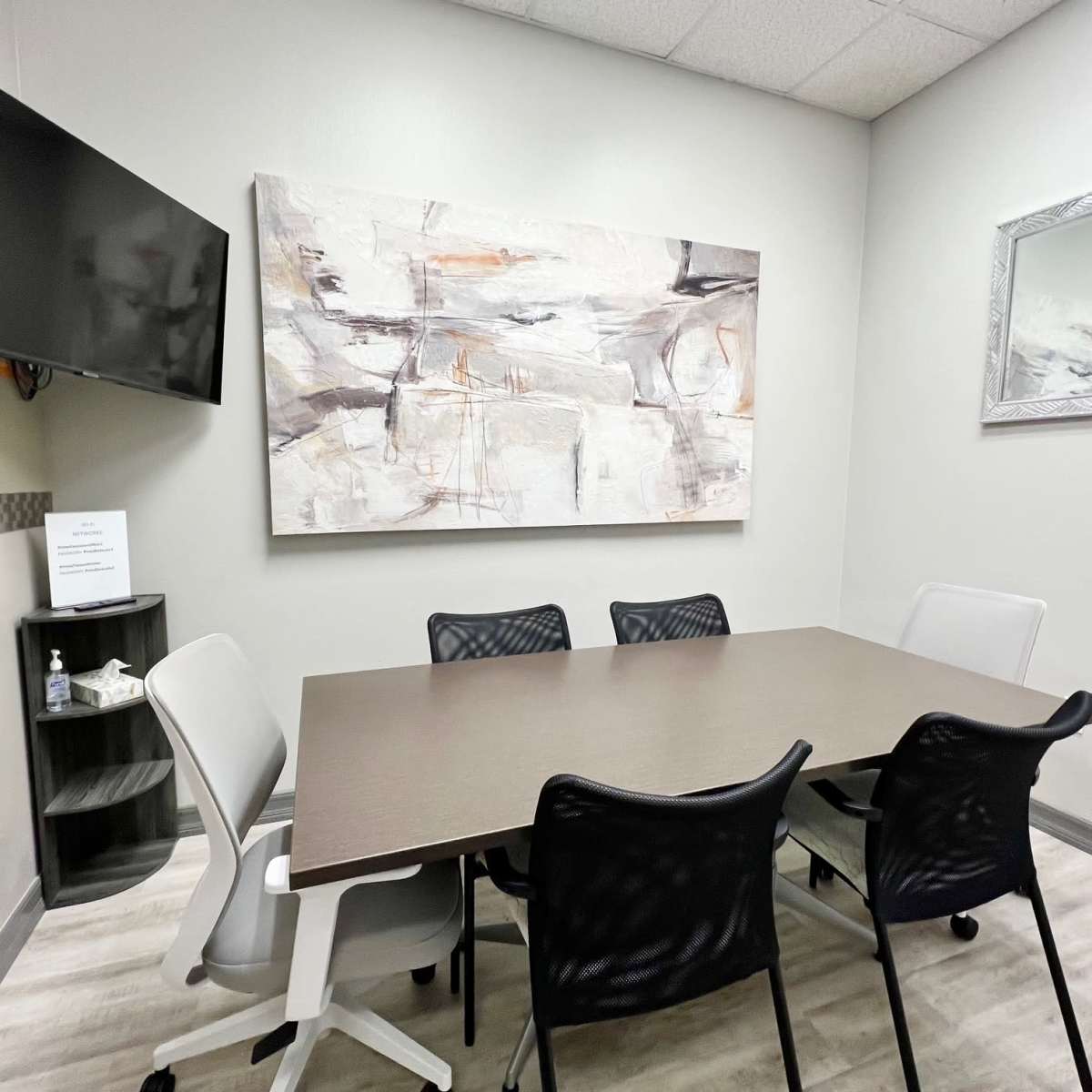 A small conference room features a rectangular table surrounded by six chairs, a wall-mounted TV, and abstract artwork.