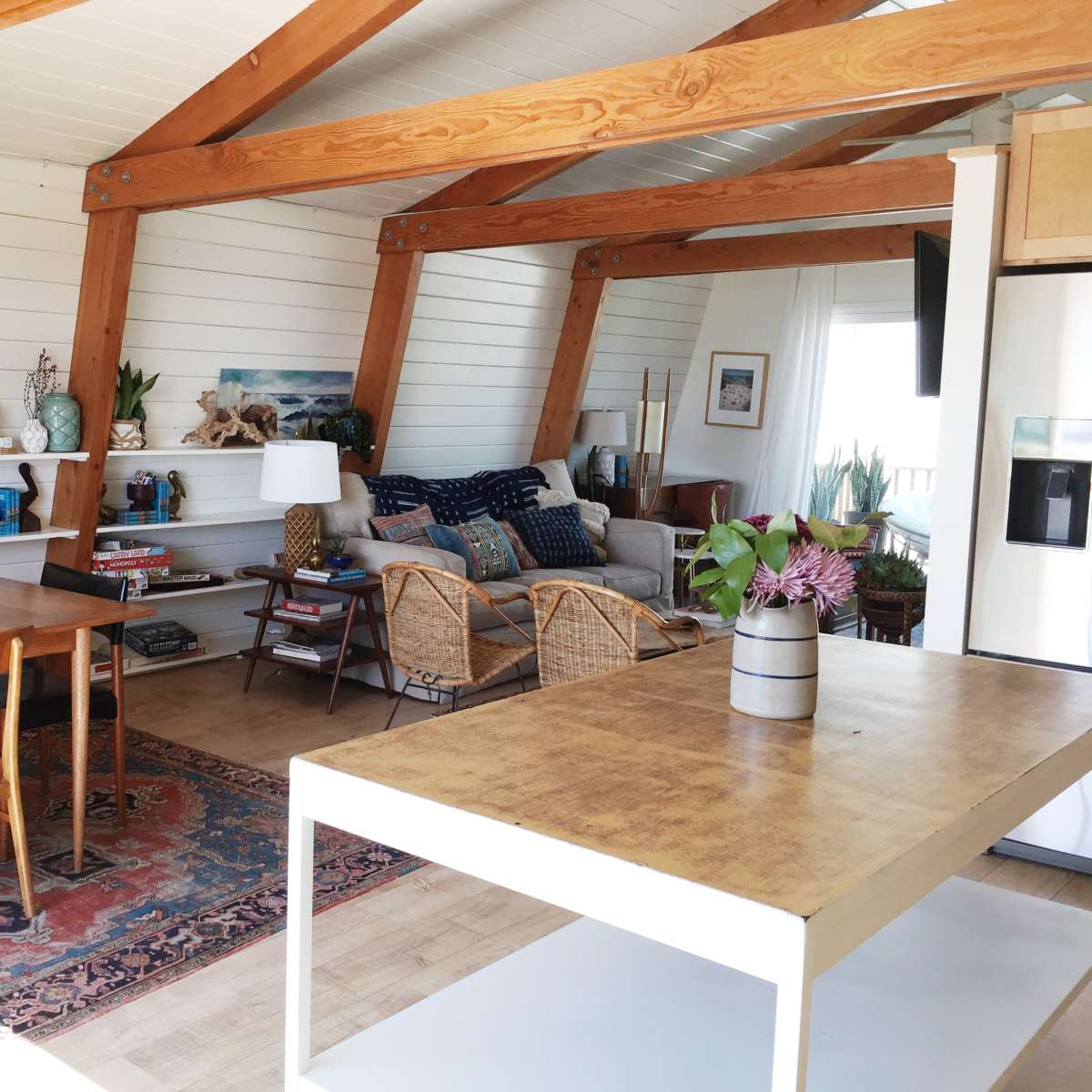 A bright and airy interior of a home with a dining table, a living area with a sofa and chairs, and wooden beams supporting the ceiling.