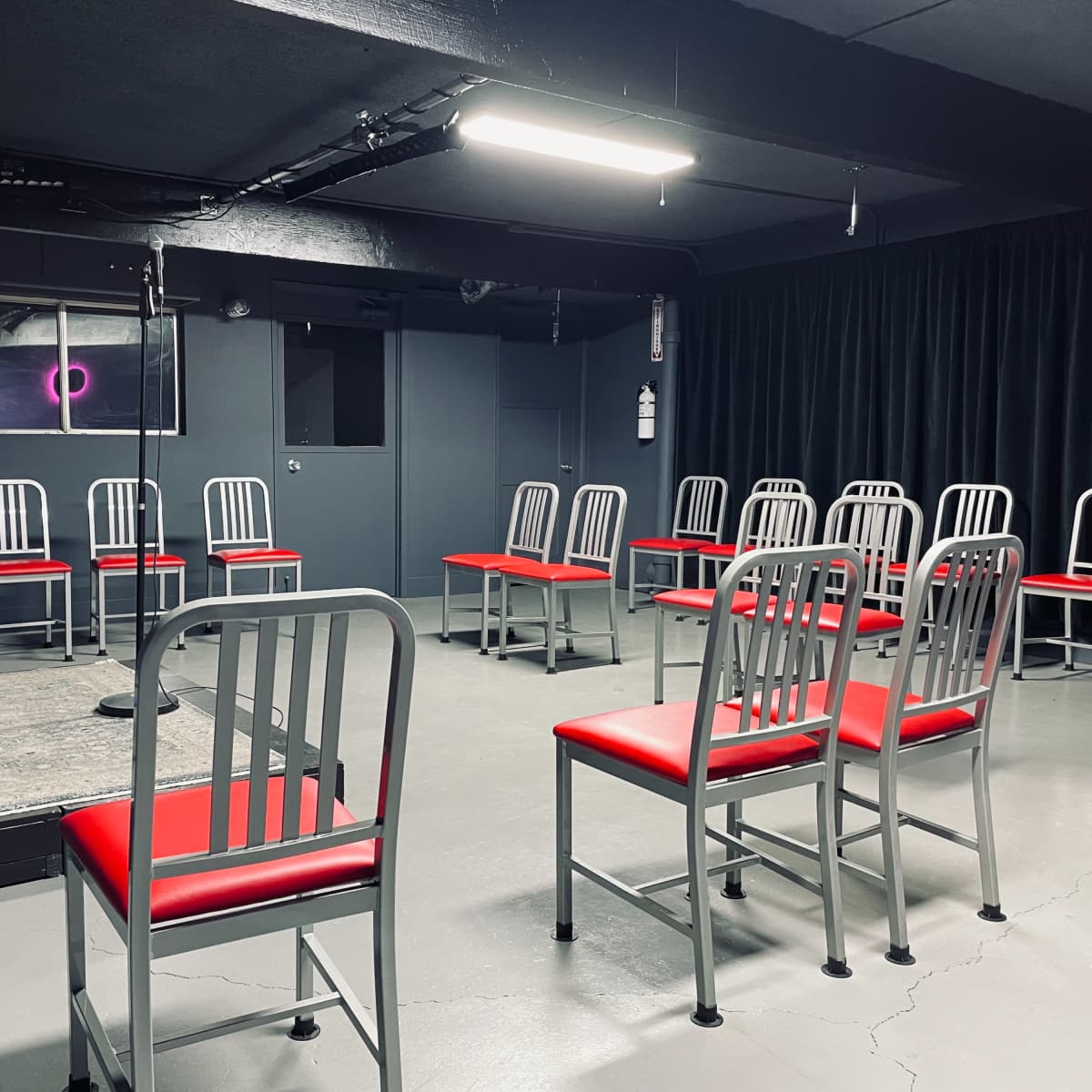 A small performance space features a blue stool and a microphone in front of a circle of red and silver chairs.