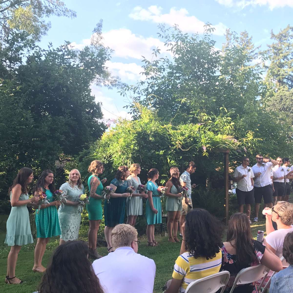 A group of women and men stand together under a trellis in a garden, while an audience sits in chairs facing them.