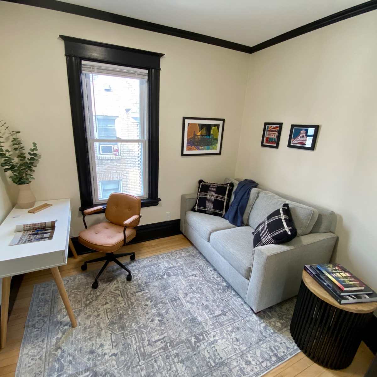 A small office space features a desk with a chair, a couch, and framed artworks on the walls.