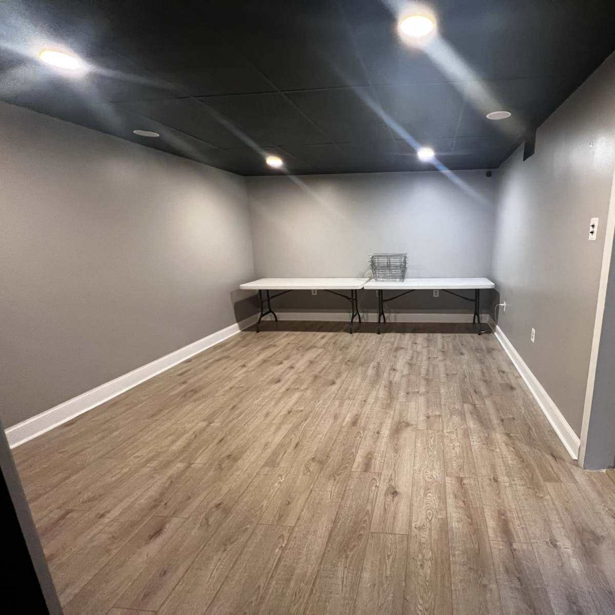 A vacant room with gray walls, wooden flooring, and two folding tables in the center under overhead lighting.