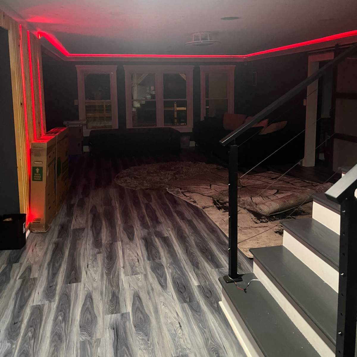 A dimly lit room with modern flooring, featuring red LED strip lighting along the walls and a staircase leading to an upper level.