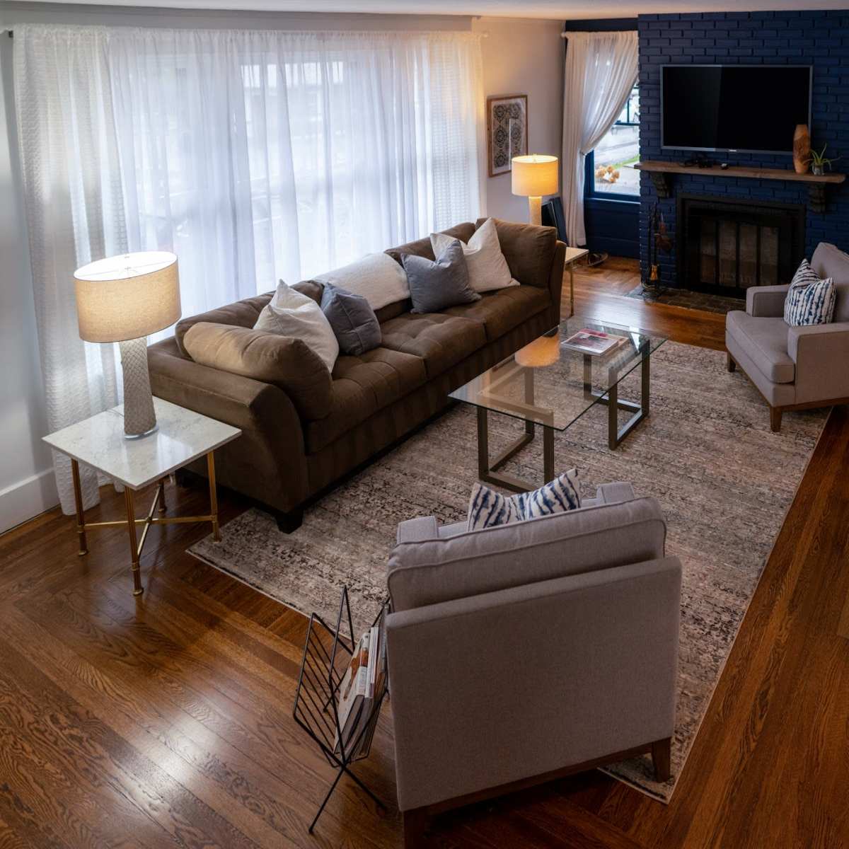 The living room features a brown sectional sofa, two light gray armchairs, a glass coffee table, and a rug on hardwood flooring, with large windows allowing natural light.