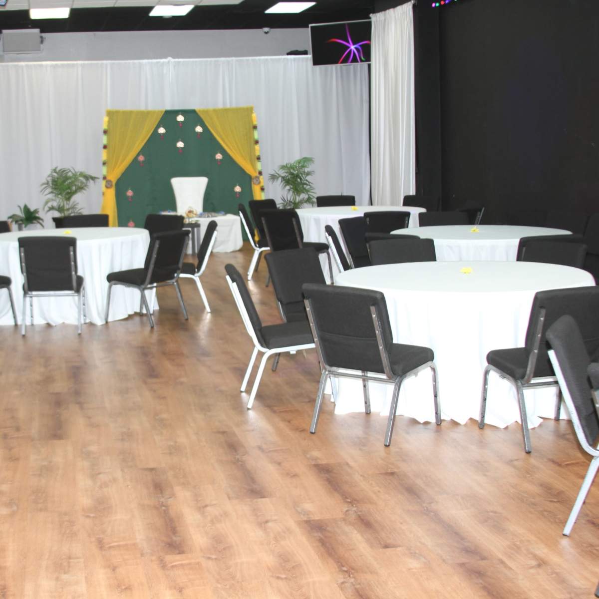 A spacious event room with round tables covered in white tablecloths and black chairs, alongside a backdrop decorated with plants and drapes.