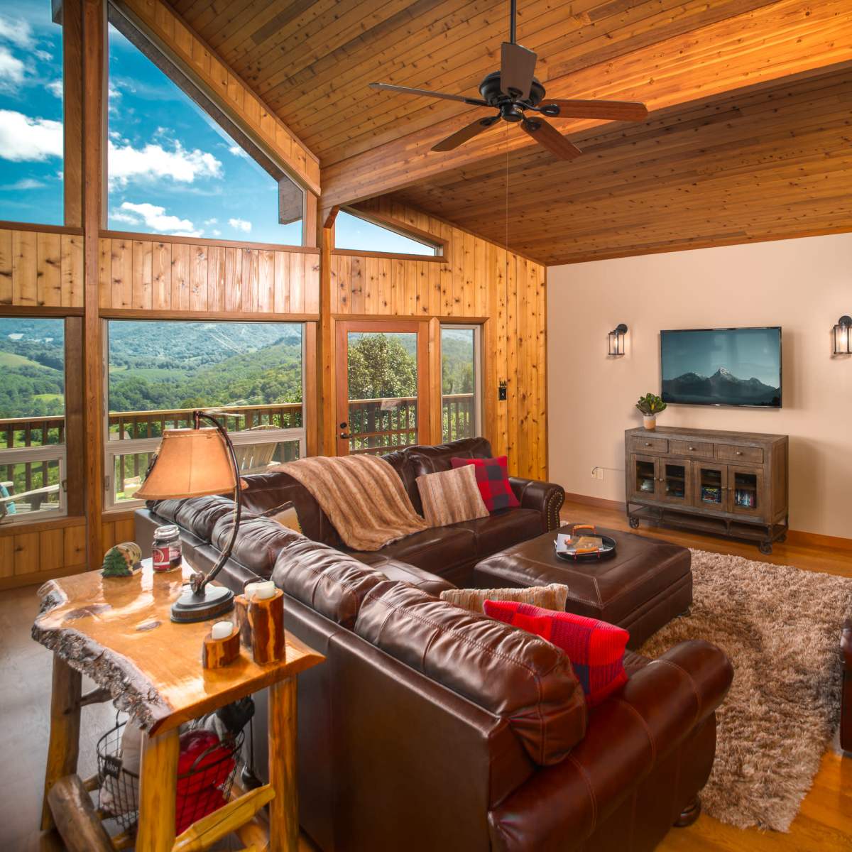 The living room features a cozy seating area with leather sofas, a wooden ceiling, and large windows offering a view of the surrounding landscape.