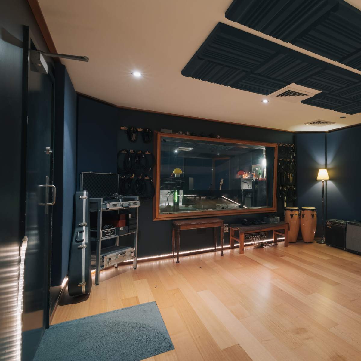 A sound studio featuring wooden floors, dark blue acoustic panels on the walls, a control window, and various musical instruments along with seating.