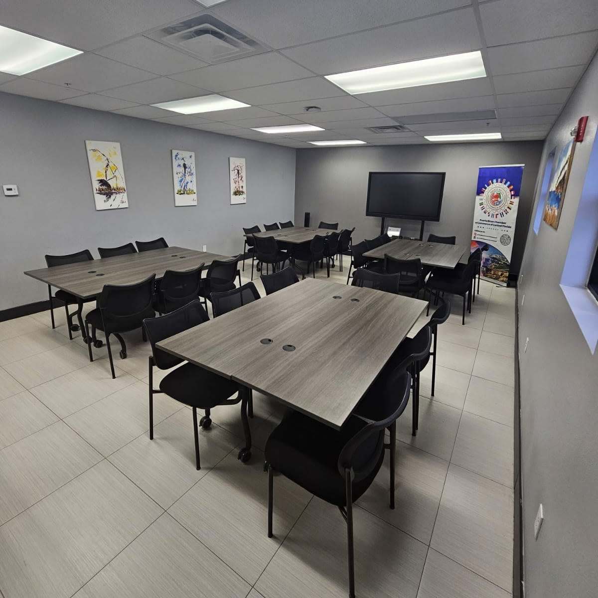 A conference room with multiple rectangular tables and black chairs arranged around them, alongside a large screen and decorative wall posters.