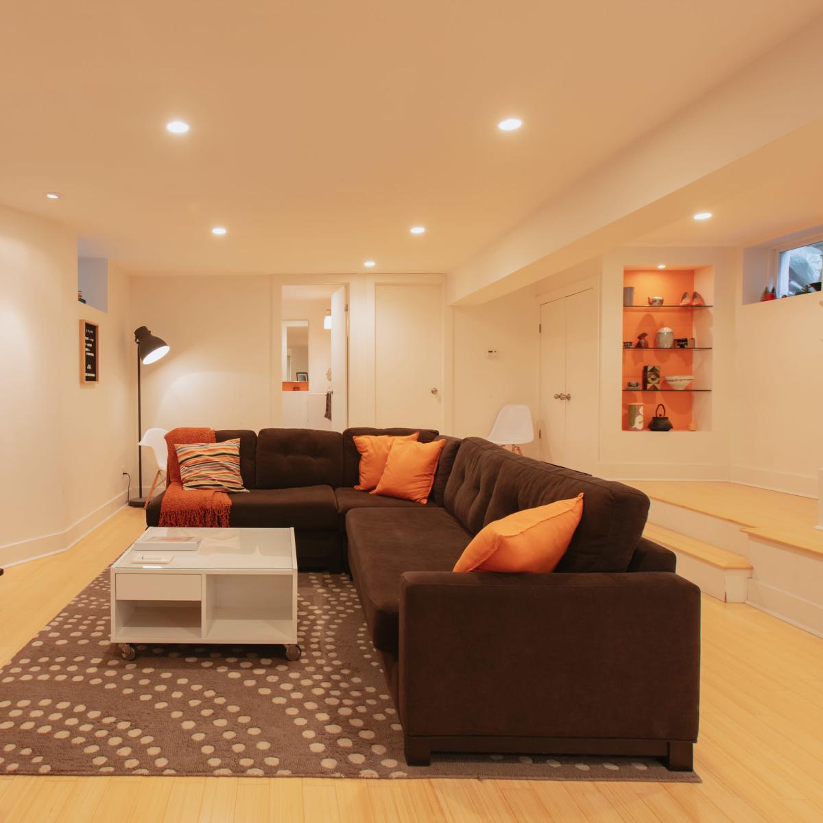 A well-lit basement living room featuring a dark sectional sofa with orange pillows, a white coffee table, a flat-screen TV, and a light-colored staircase.