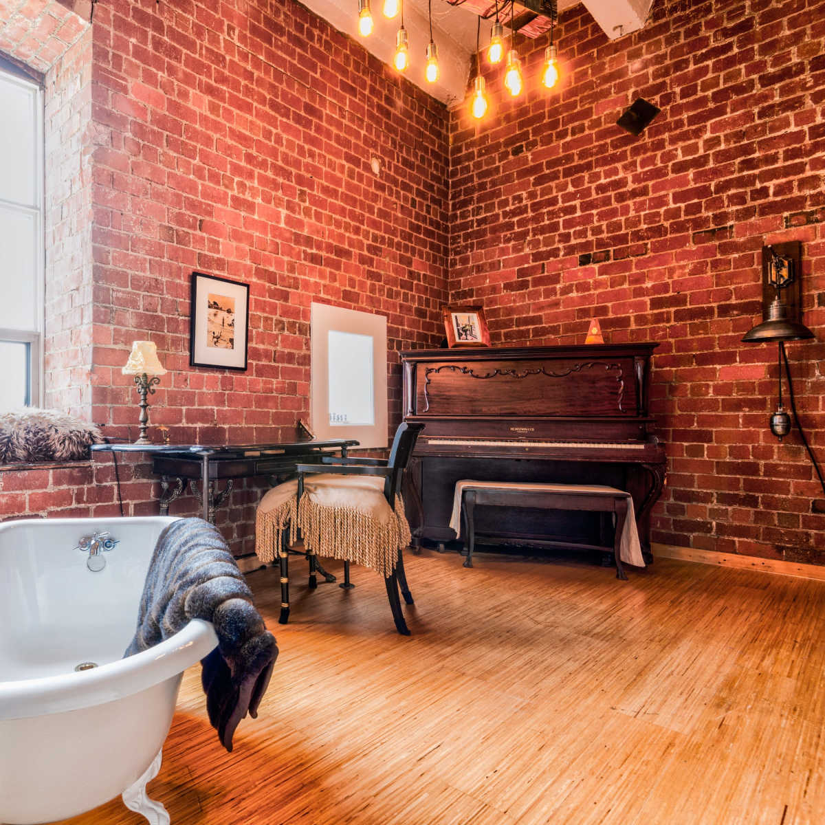 A vintage-style room featuring a clawfoot bathtub, a piano, a table with a chair, and exposed brick walls illuminated by pendant lights.