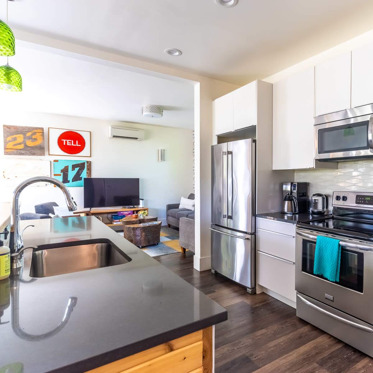 A modern kitchen features stainless steel appliances, a dark countertop, and a view of a living area with a gray sofa and colorful decor.