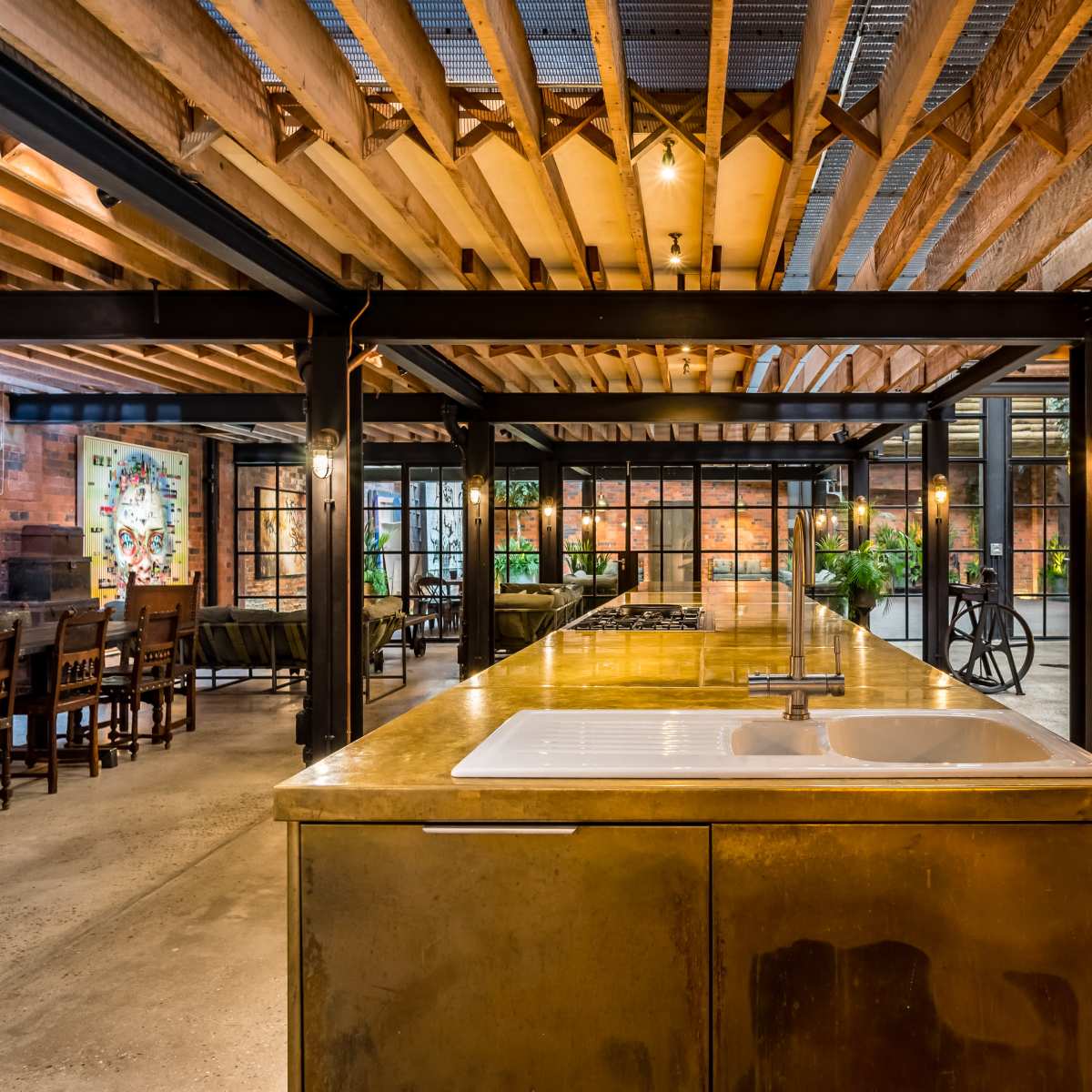 A modern industrial kitchen with a large metal countertop and exposed wooden ceiling beams, surrounded by a spacious dining area with brick walls and large windows.