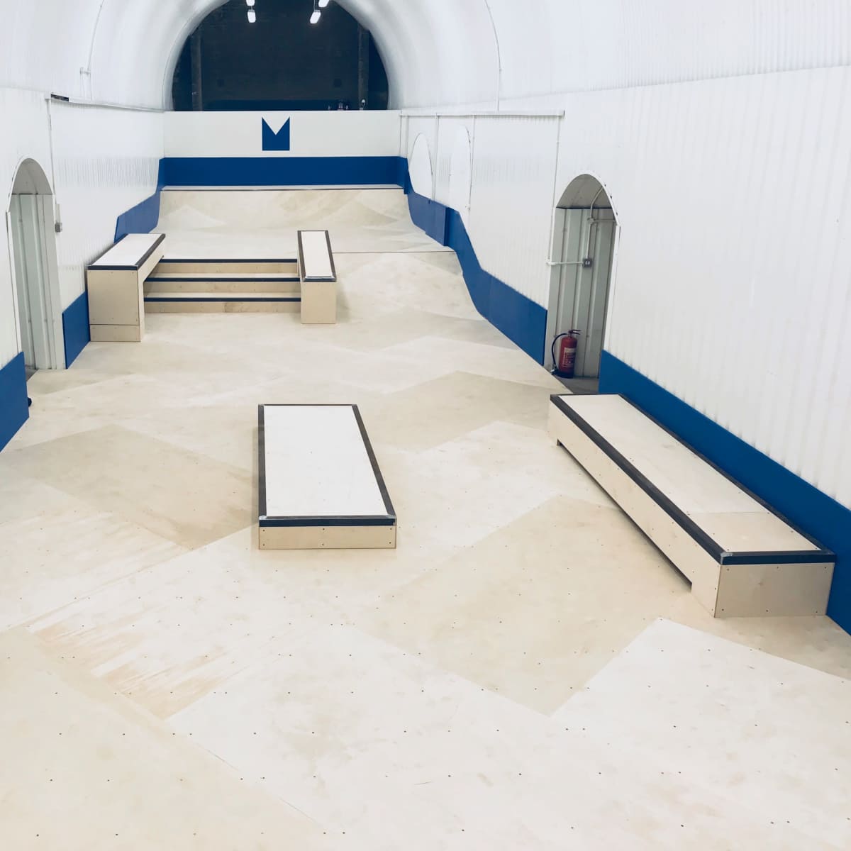 A skatepark interior featuring multiple ramps and flat surfaces arranged in a tunnel-like structure with white walls and blue accents.