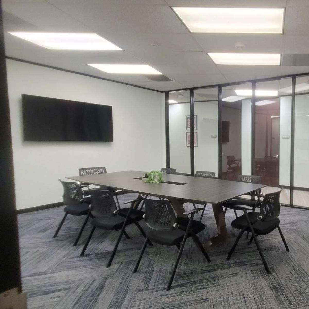A modern conference room features a large rectangular table surrounded by black chairs, with glass walls and a television mounted on one side.