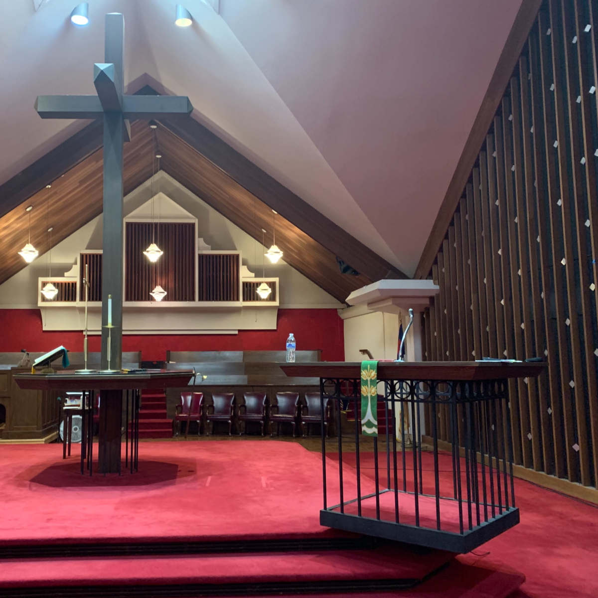 The interior of a church features a large cross at the center of a raised altar area, surrounded by pews and decorative elements against a backdrop of wooden architecture.
