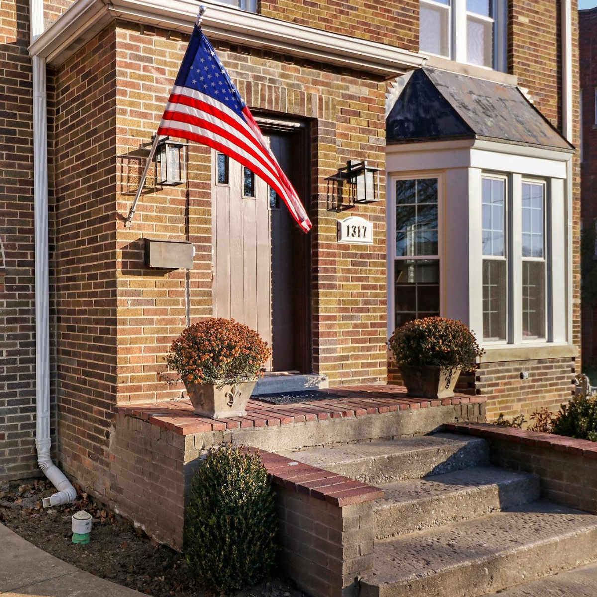 A brick house with a staircase leading to the front door, featuring an American flag hanging from the porch and potted plants on either side of the entrance.