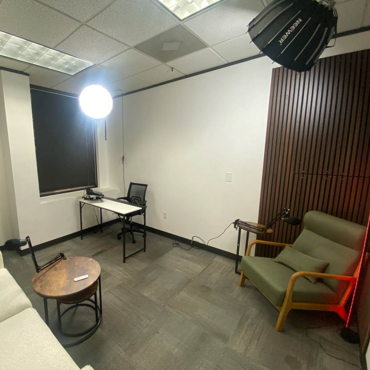 A small, sparsely furnished office space featuring two seating chairs, a coffee table, and a desk with a chair, illuminated by a large light source.