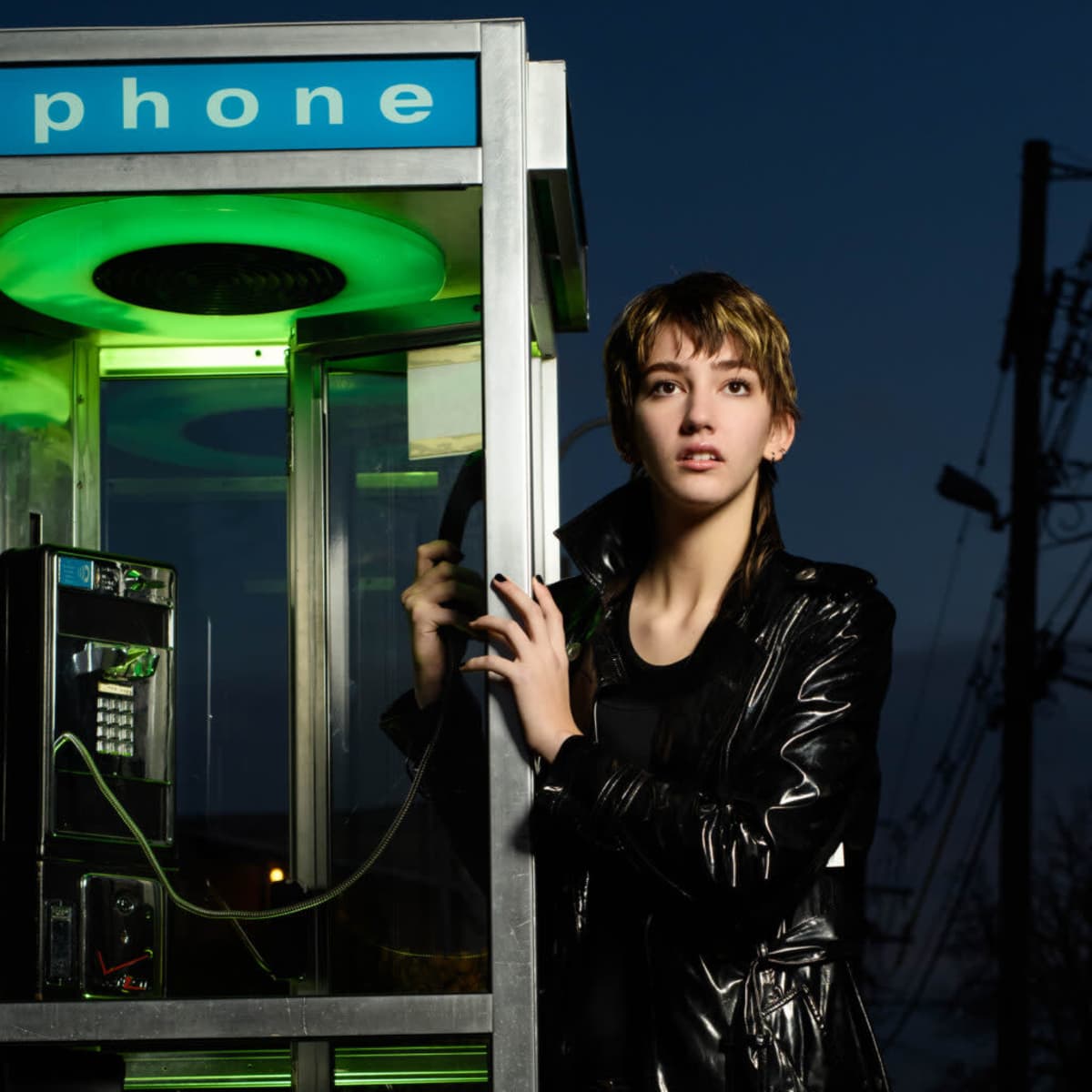 A person stands beside a brightly lit phone booth at dusk, wearing a shiny black coat.