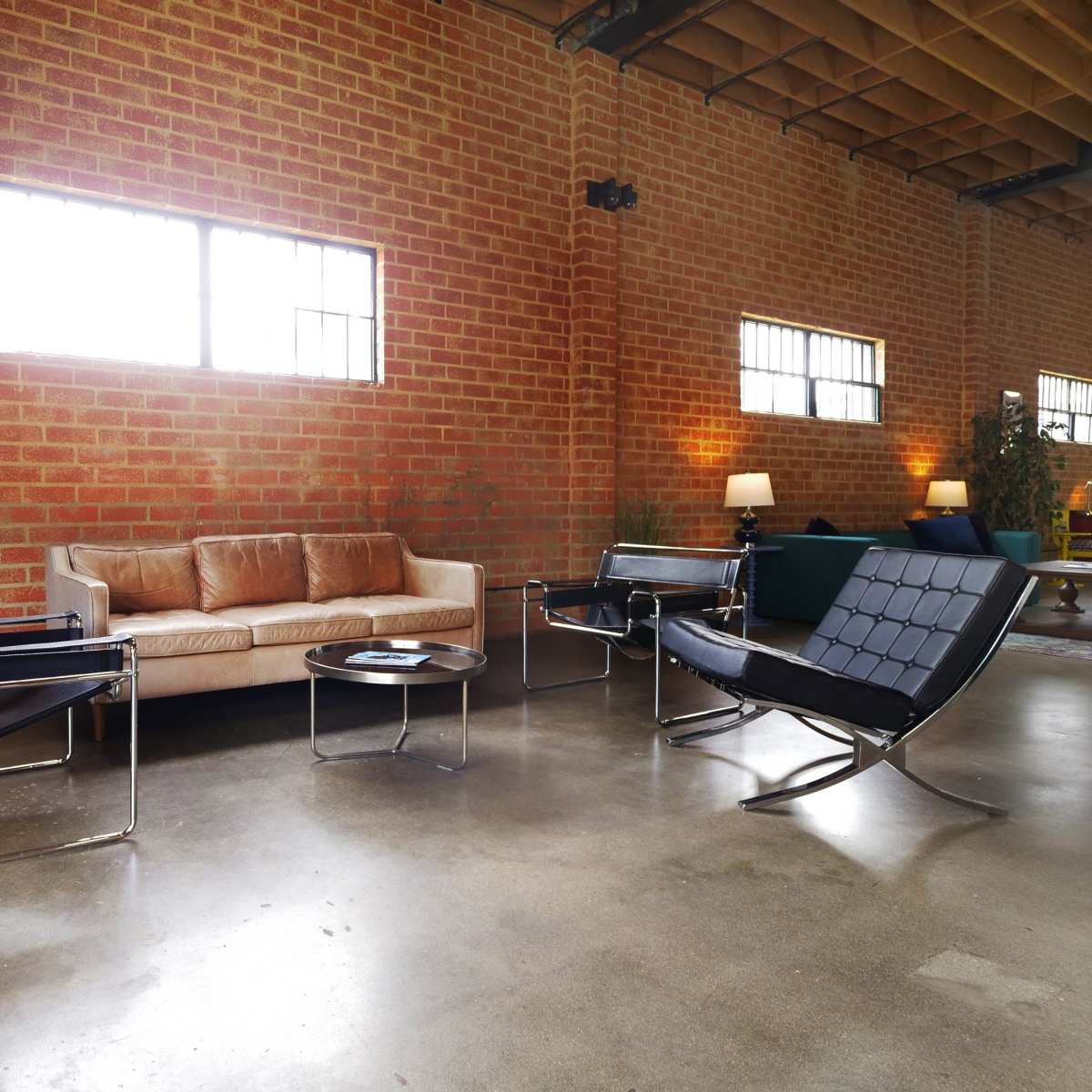 The interior features a spacious room with exposed brick walls, a couch, two modern chairs, a coffee table, and large windows letting in natural light.