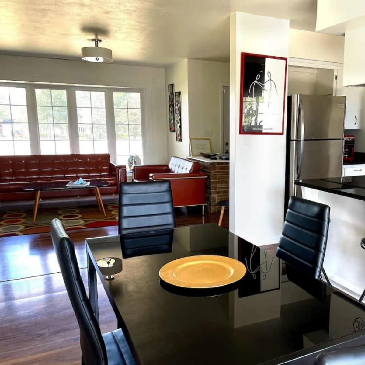 A modern open-concept living and dining area with a glass dining table and black chairs, adjacent to a kitchen featuring stainless steel appliances and a red sofa in the background.